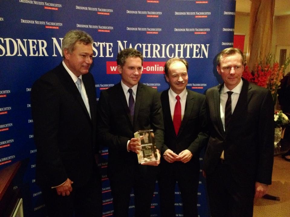 Four men in suits stand together at an event, with one holding a trophy, in front of a backdrop featuring the "Dresdner Neueste Nachrichten" logo.