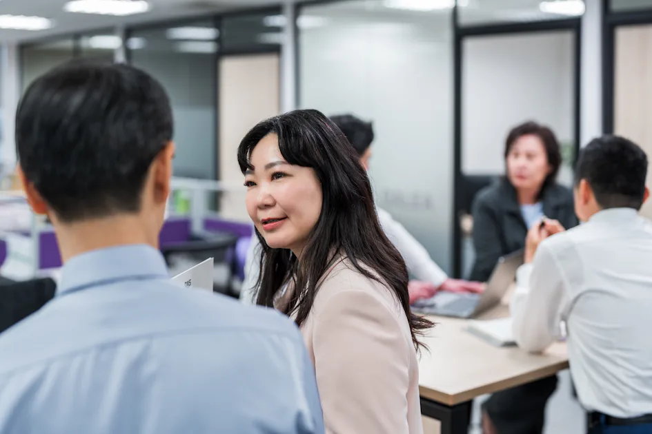 Group meeting 辦公室場景中,幾位專業人士坐在桌子旁交談,背景可見玻璃隔間和文件,這營造出一種協作工作環境的氛圍。