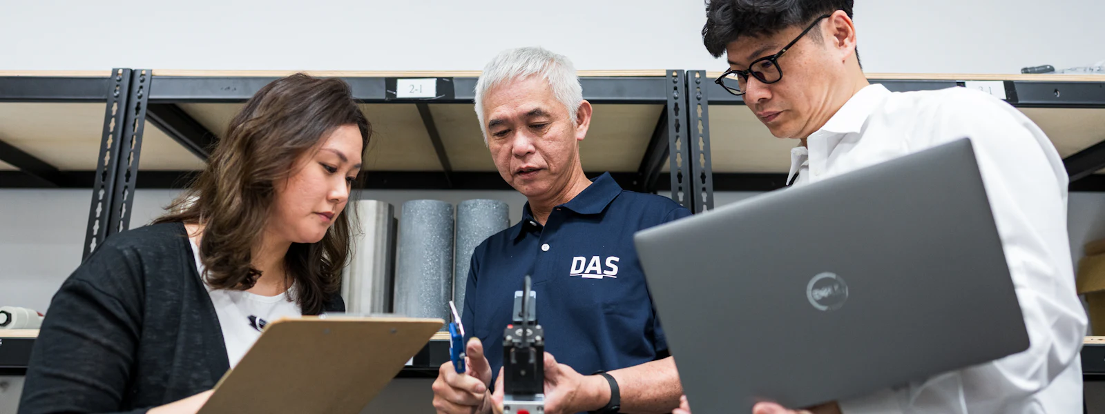 料件討論 三人在辦公室內進行討論,其中一人持有筆記本電腦,另一人拿著文件夾,第三人在講解機械裝置。