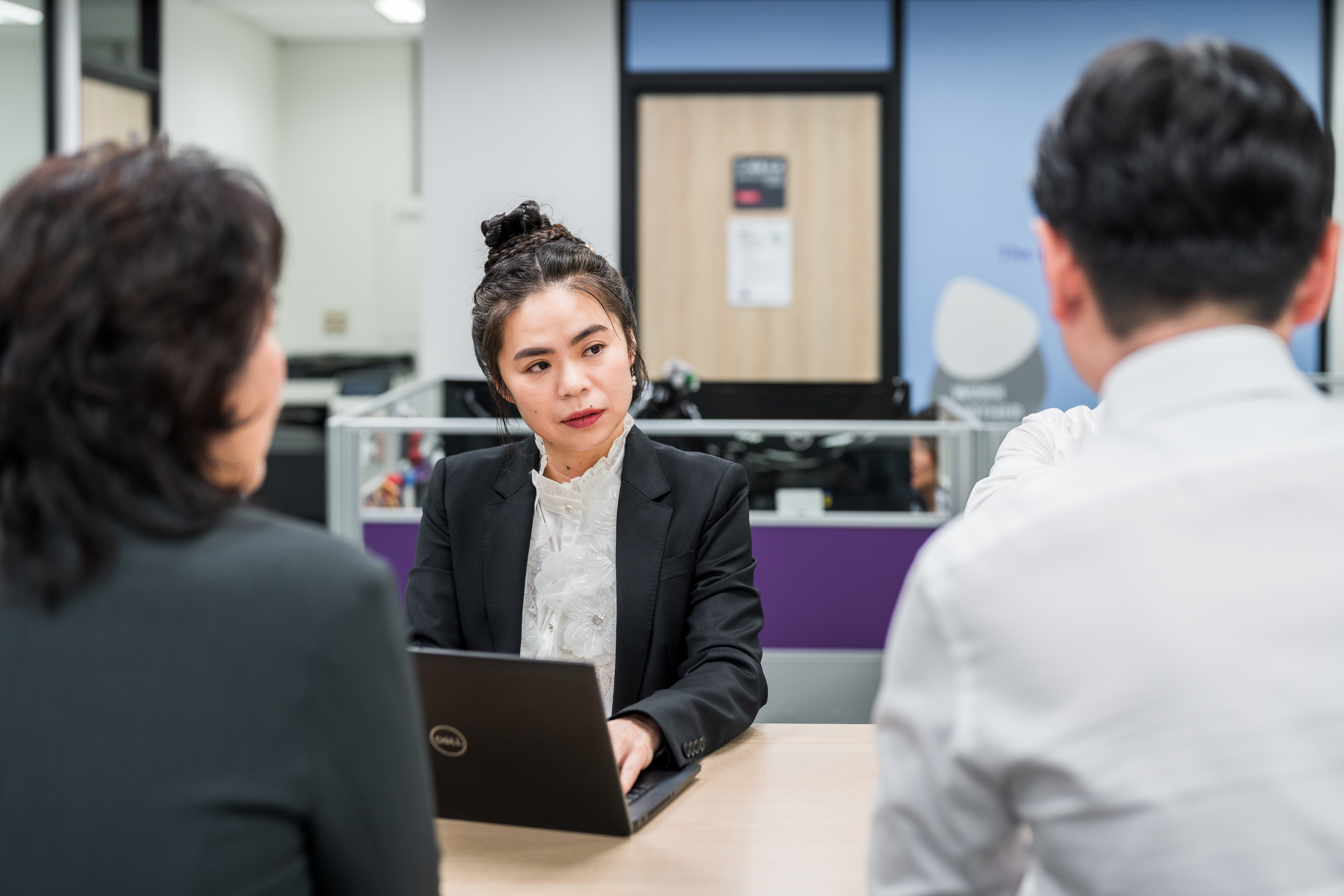 三位商務人士在現代辦公室會議桌旁進行討論，中間的人手持筆記型電腦，表情專注。
