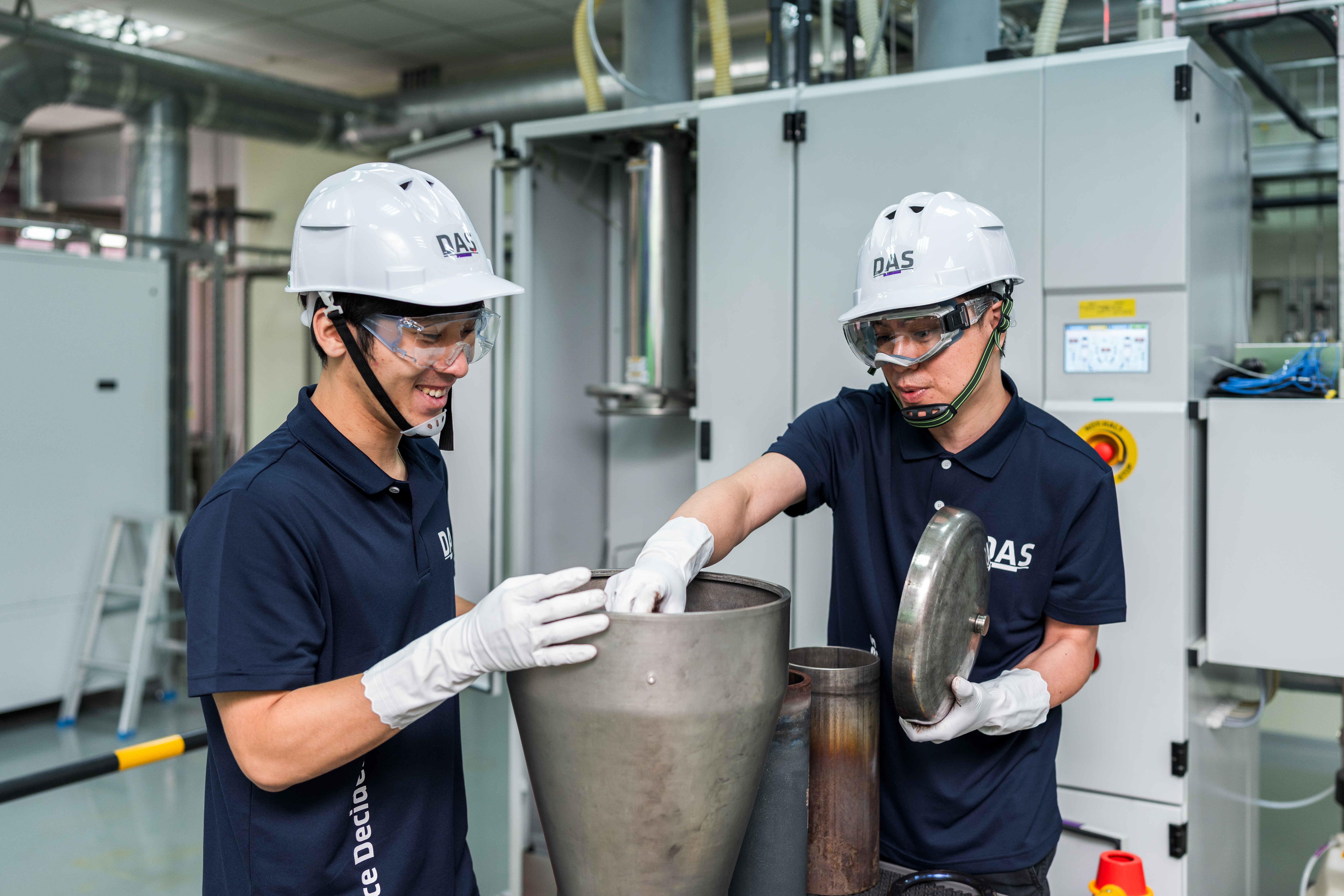 兩名穿著深色工作服和白色安全帽的工人正在工廠內合作操作大型金屬設備，專注於將一個金屬蓋放回設備上，背景是現代化的工業設施。