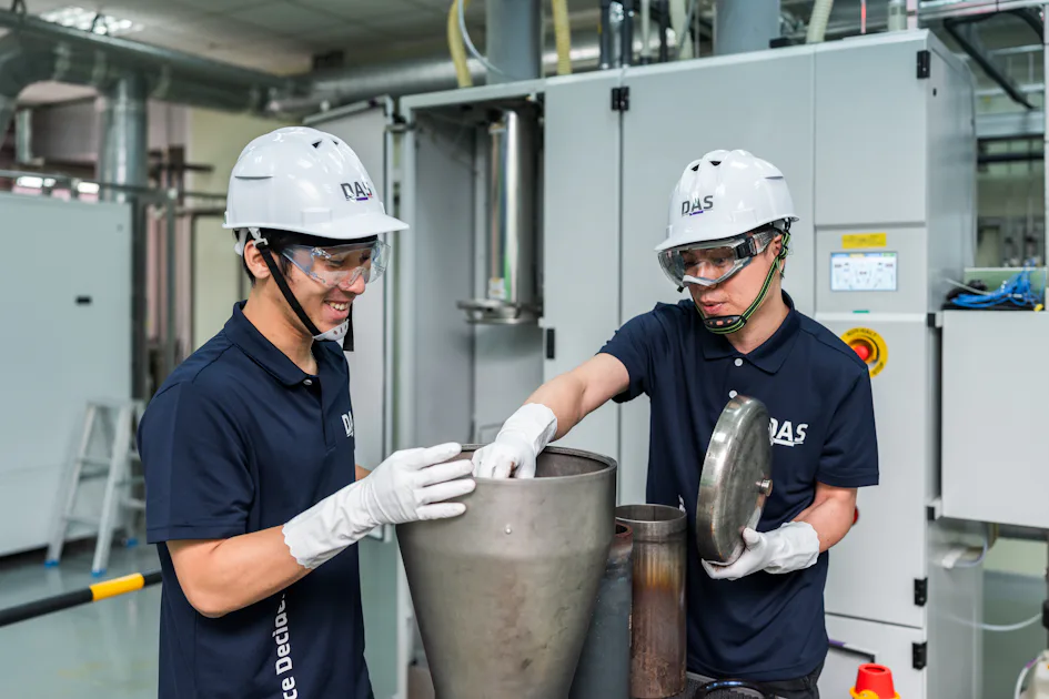 parts 確認 兩名穿著深色工作服和白色安全帽的工人正在工廠內合作操作大型金屬設備,專注於將一個金屬蓋放回設備上,背景是現代化的工業設施。