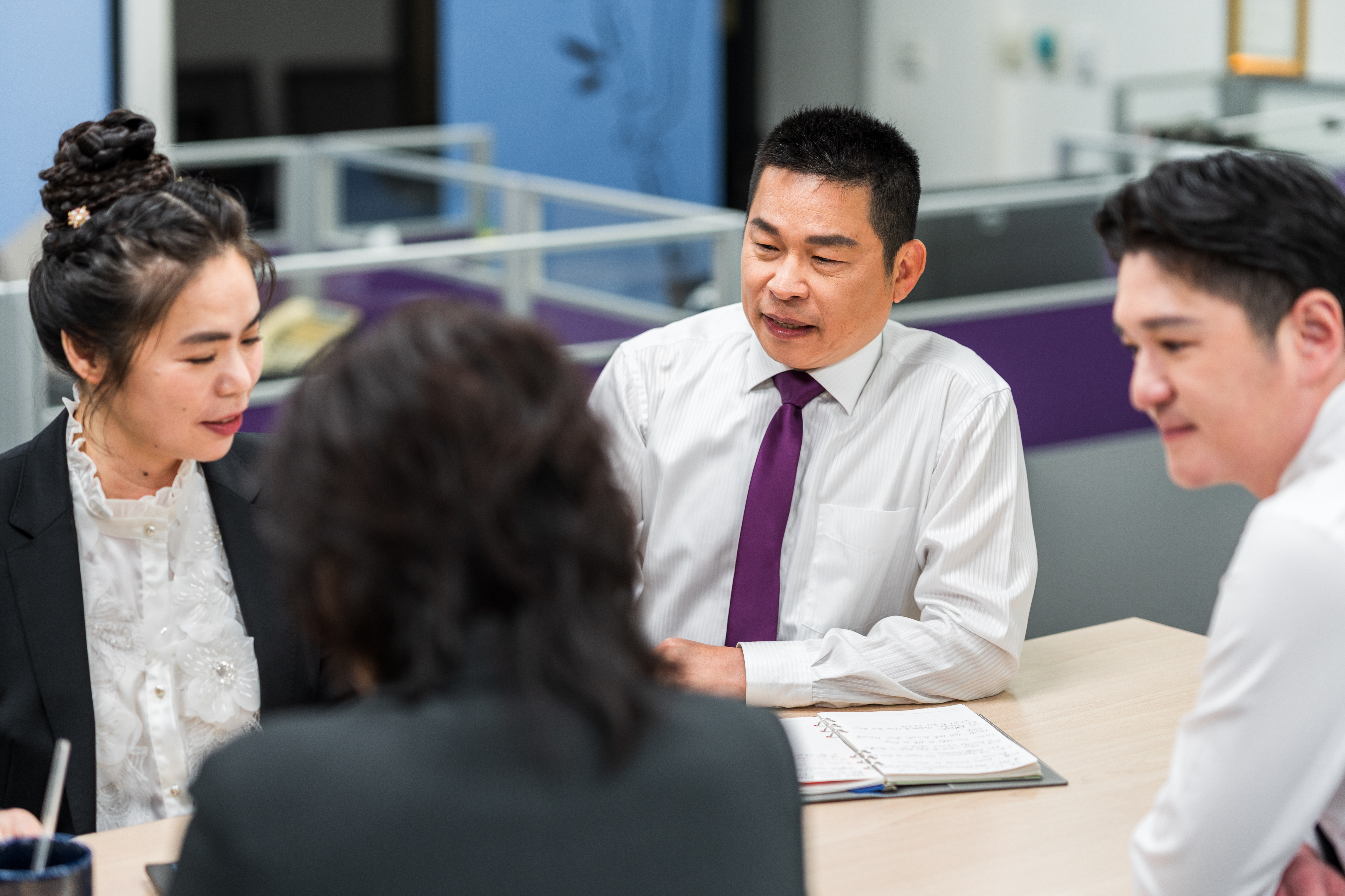 四個人坐在會議桌旁開會，三位身穿正式商務服裝的人正專注地進行討論，桌上擺有筆記本和筆。