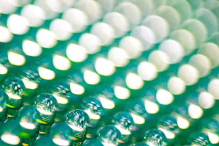 LED industry The image displays a close-up of a textured surface with numerous small, round, reflective droplets arranged in rows, reflecting light in a pattern that creates a shimmering, blurred, green and white bokeh effect.