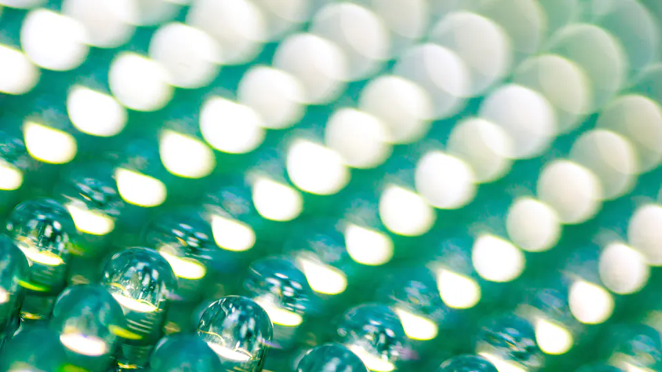 LED industry The image displays a close-up of a textured surface with numerous small, round, reflective droplets arranged in rows, reflecting light in a pattern that creates a shimmering, blurred, green and white bokeh effect.