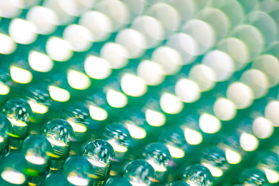 The image displays a close-up of a textured surface with numerous small, round, reflective droplets arranged in rows, reflecting light in a pattern that creates a shimmering, blurred, green and white bokeh effect.