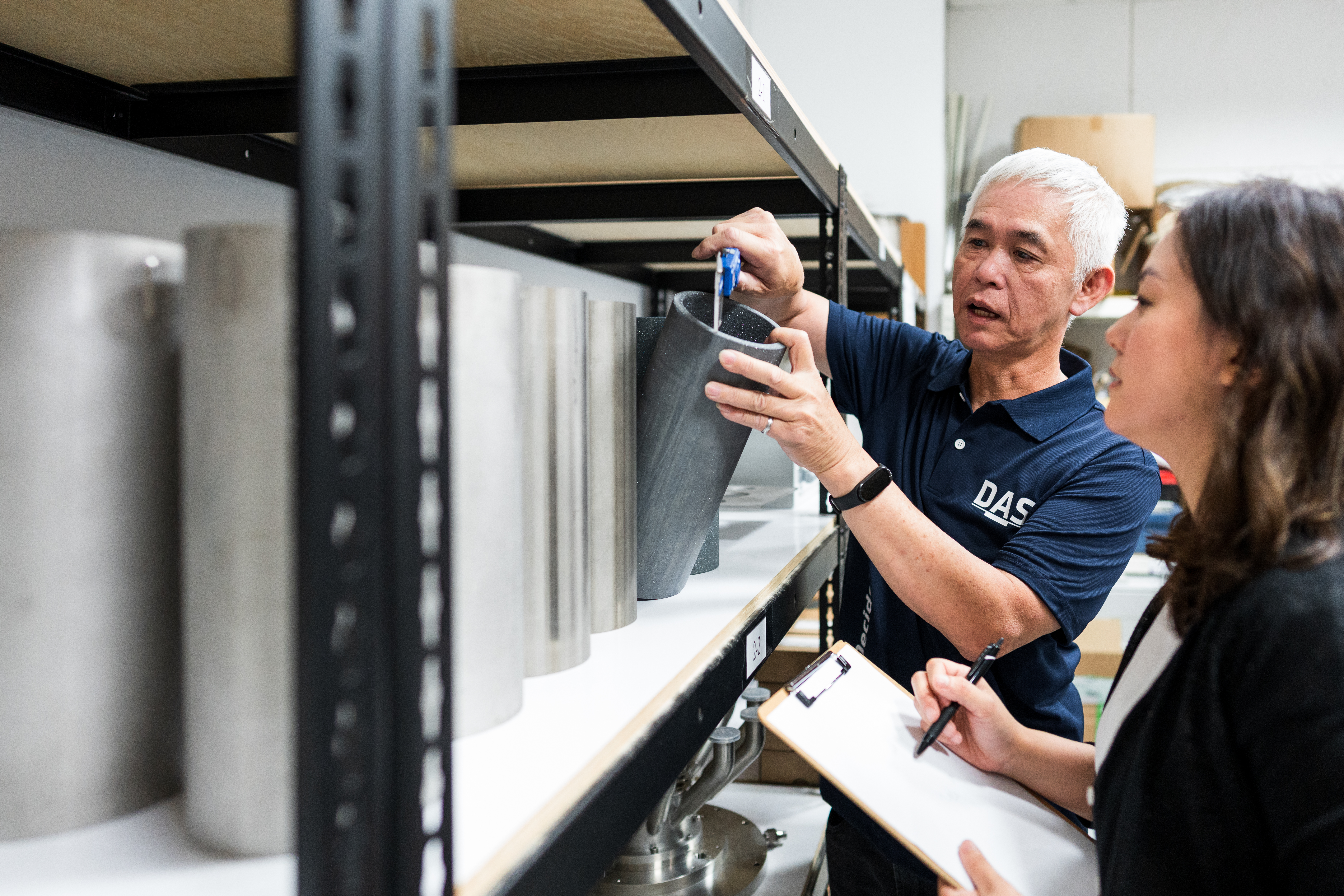 兩位工作人員在倉庫中檢查一個大型金屬零件，其中一人手持計量工具，另一人正在筆記本上記錄數據。