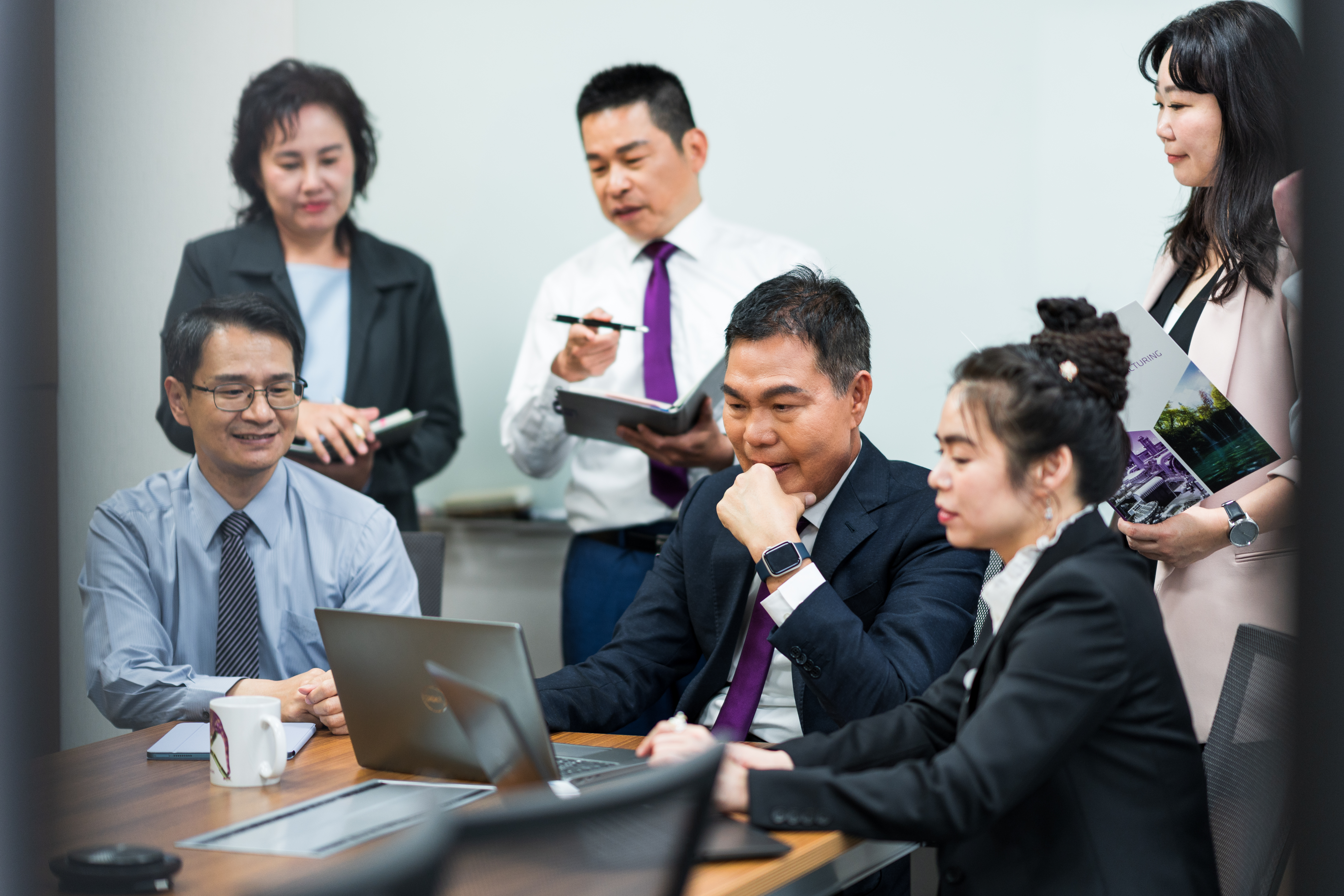 一群商務人士在會議室中專注於筆記型電腦屏幕，進行團隊合作和交流，幾位成員正忙於記錄和討論。