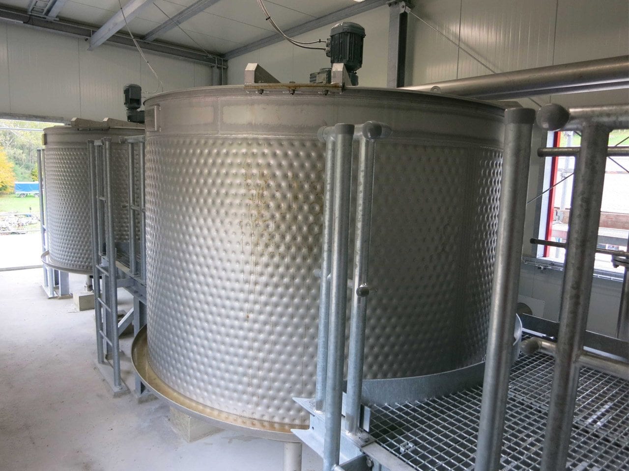 Large stainless steel fermentation tanks are lined up in an industrial winery setting, showcasing the robust and modern equipment used for winemaking.