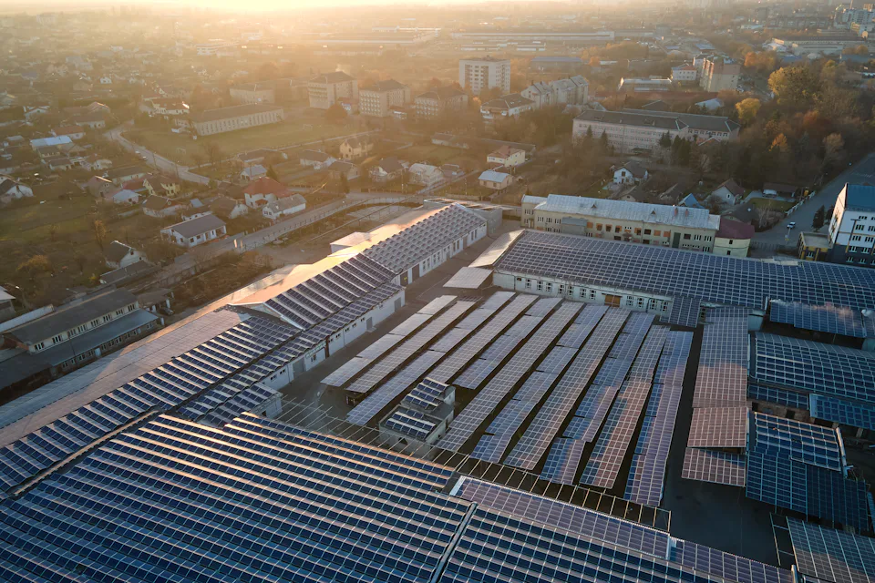 solar industry Aerial view of an expansive solar panel farm on the rooftops of industrial buildings in an urban area at sunset, showcasing sustainable energy solutions amidst a sprawling cityscape.