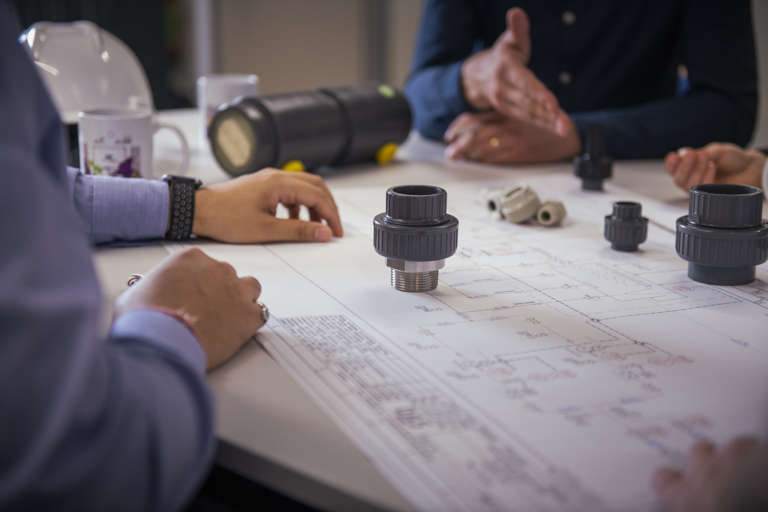 A group of people collaborates over a large technical blueprint spread on a table, with various pipe fittings and connectors placed on the plan, indicating a discussion or planning session related to plumbing or engineering design.
