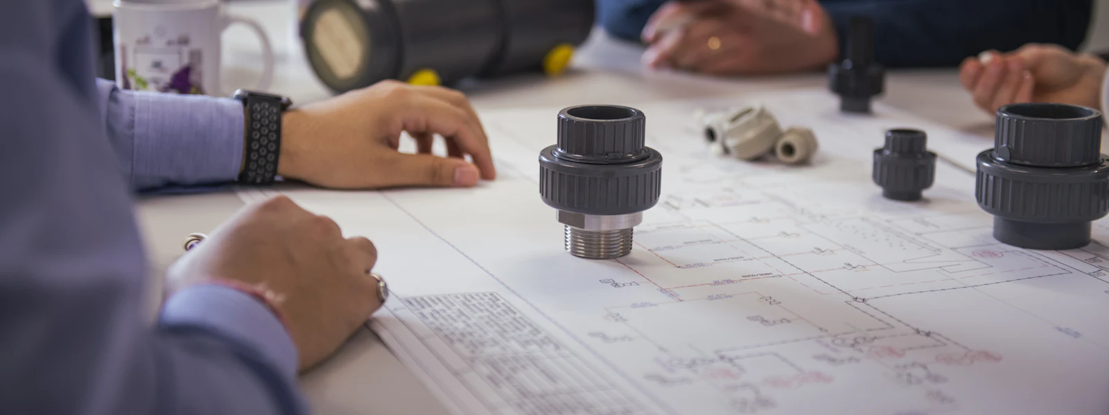 기술 구성 요소 이미지 여러 사람이 테이블 위에 펼쳐진 대형 기술 설계도를 바탕으로 협업하고 있으며, 다양한 파이프가 보입니다.