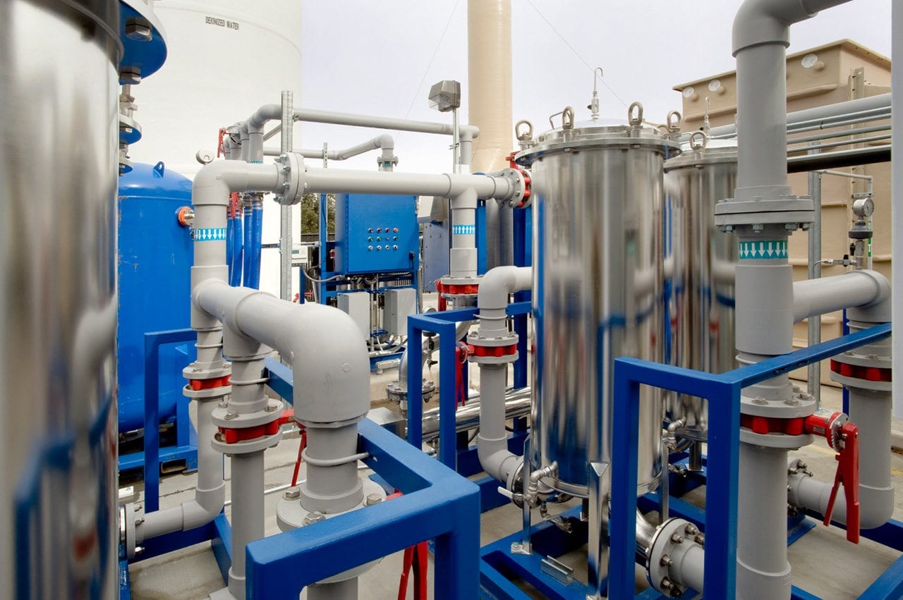 Industrial facility with interconnected shiny metal tanks and large gray pipes, featuring blue structural supports and several valves, indicating a complex water treatment or chemical processing system.