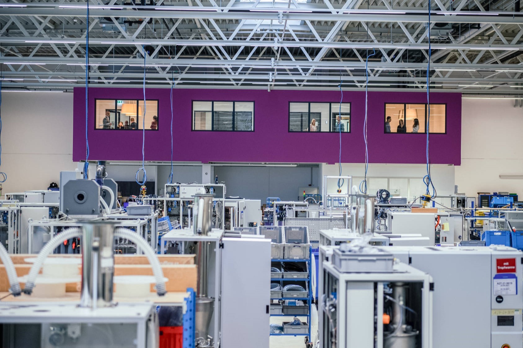 In a large, modern factory hall with numerous machines and devices, a purple wall can be seen in the background, overlooked by a raised control room with windows.
