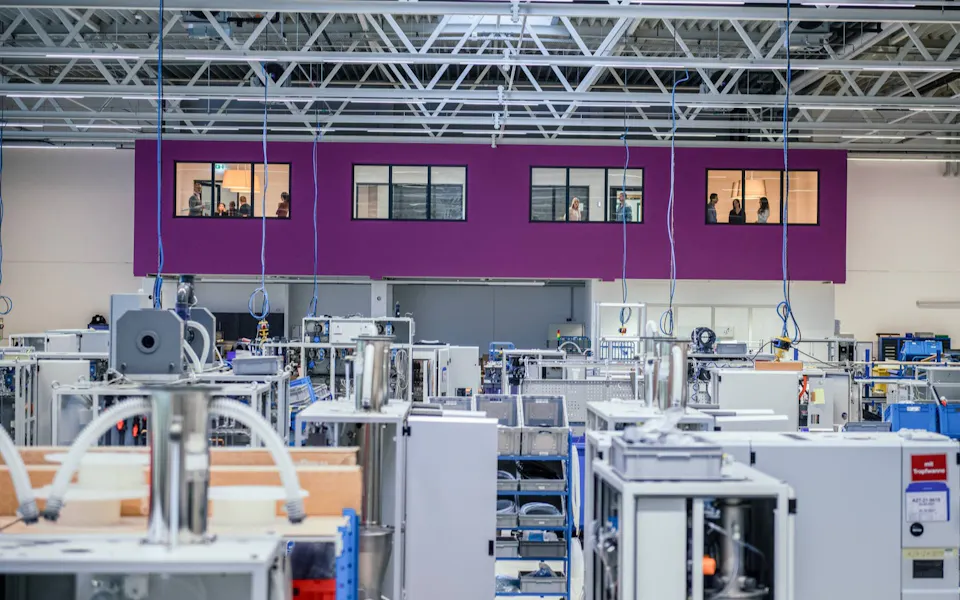 Production at DAS EE In a large, modern factory hall with numerous machines and devices, a purple wall can be seen in the background, overlooked by a raised control room with windows.