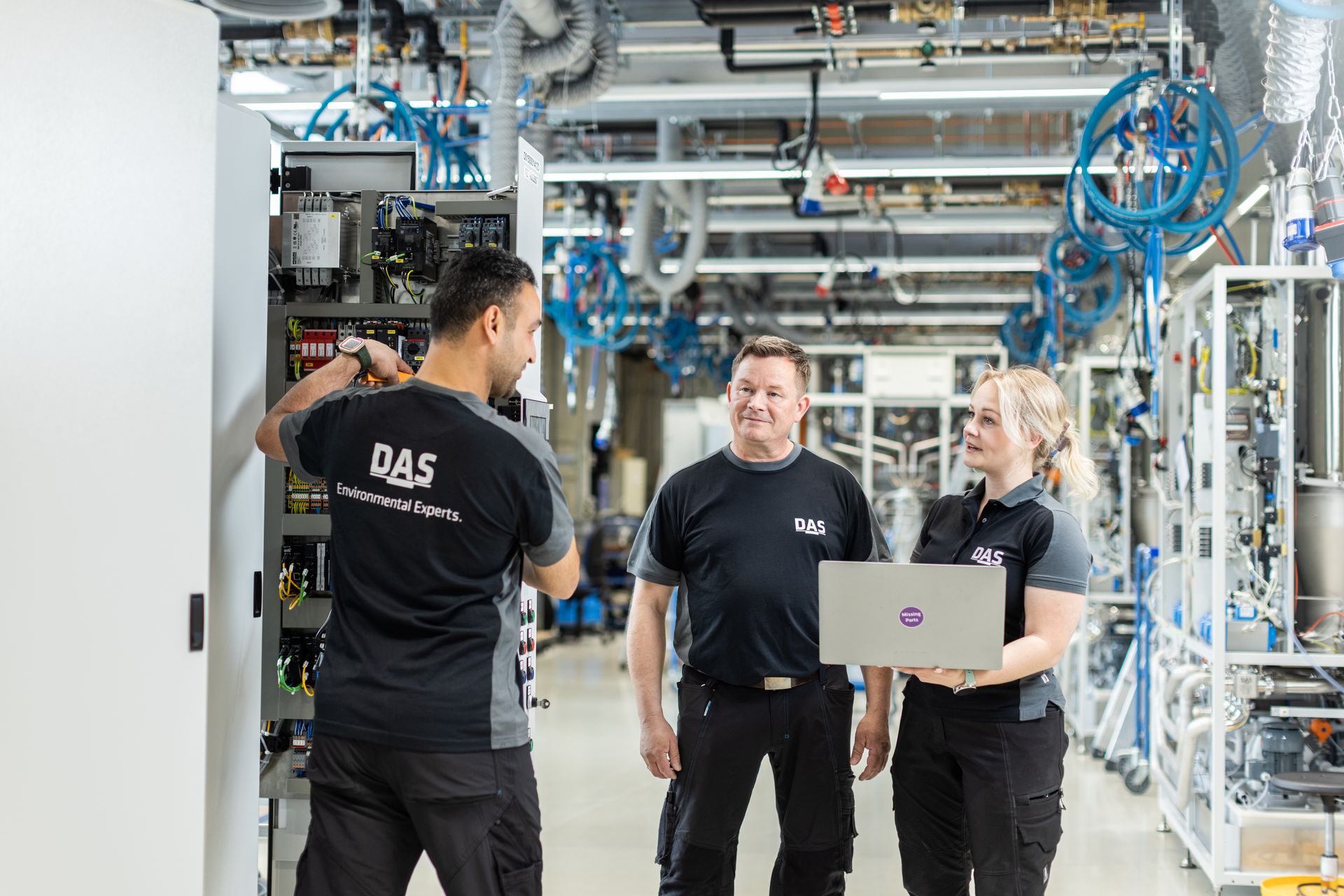 Three individuals in work clothes standing in a modern factory environment, with one person working on a control cabinet and the others standing next to it with a laptop; numerous pipes and machines can be seen in the background.