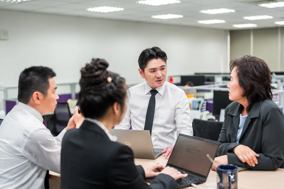 團體討論 四位商務人士在現代辦公室的會議桌旁開會,專注討論項目計畫,桌上擺放著筆記本電腦,背景為開放式辦公空間和明亮的燈光。