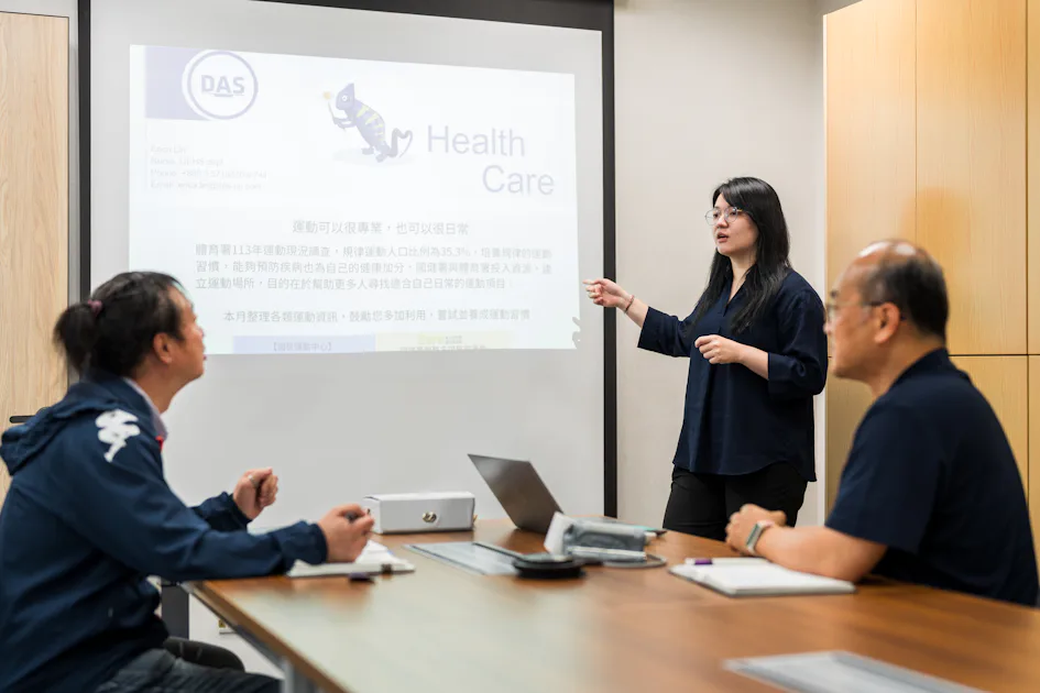 醫療簡報 會議室中,一名正在演講的女性指向螢幕上的醫療簡報,兩名坐著的男士專注地聆聽。