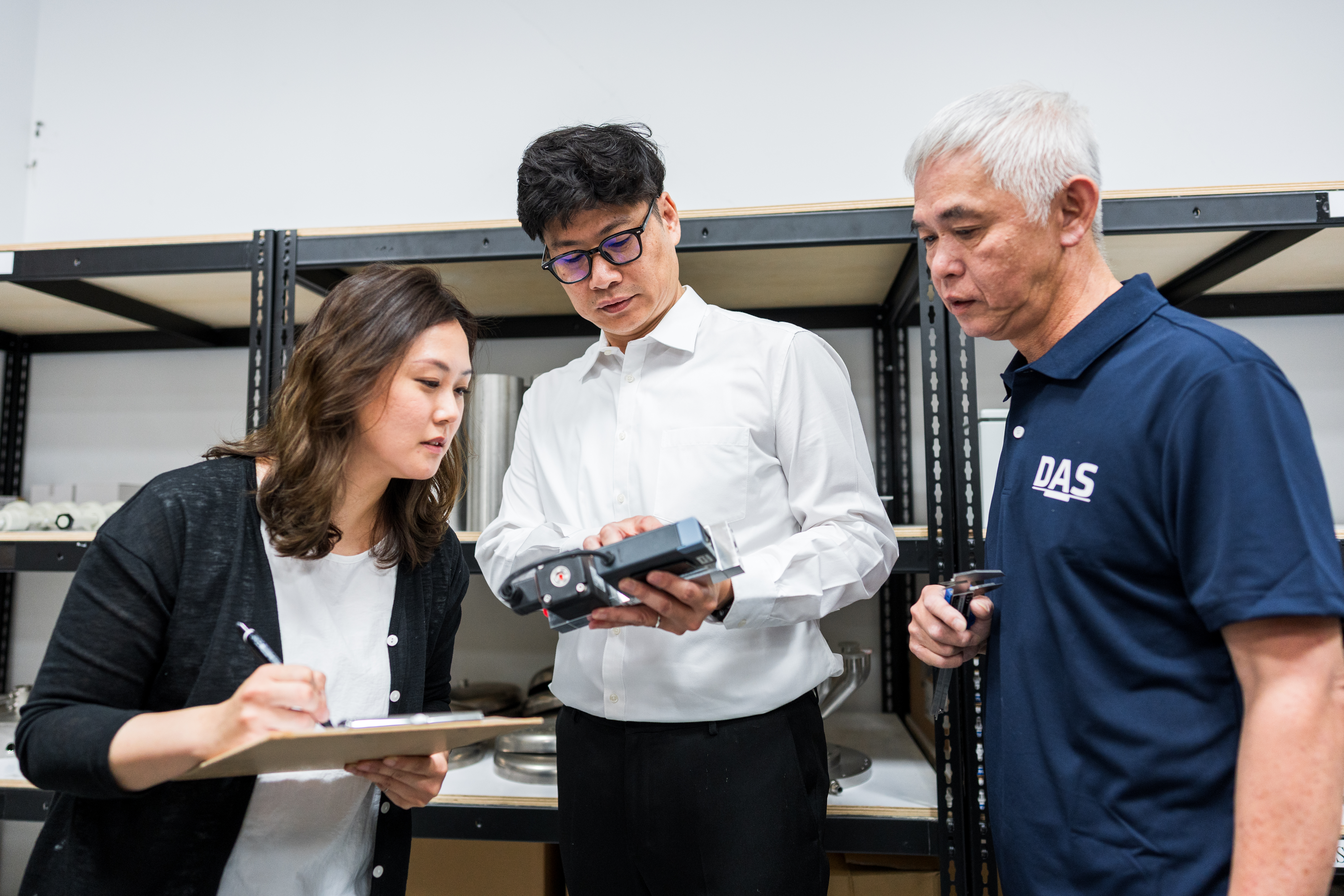 三位穿著正式服裝的人站在儲物架前，一位女性正在查看文件夾，兩位男性在討論檢查設備。
