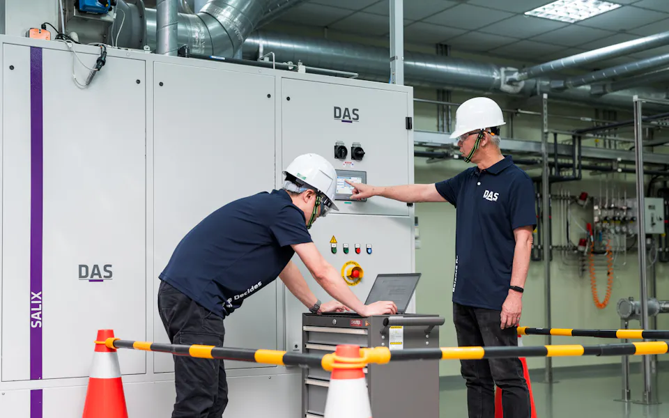 機台處理 兩位穿著深藍色制服和白色安全帽的人員在工業控制室進行設備監測,他們站在大型白色控制面板前,使用筆記型電腦檢查數據,旁邊設有安全障礙構件和反光交通錐。