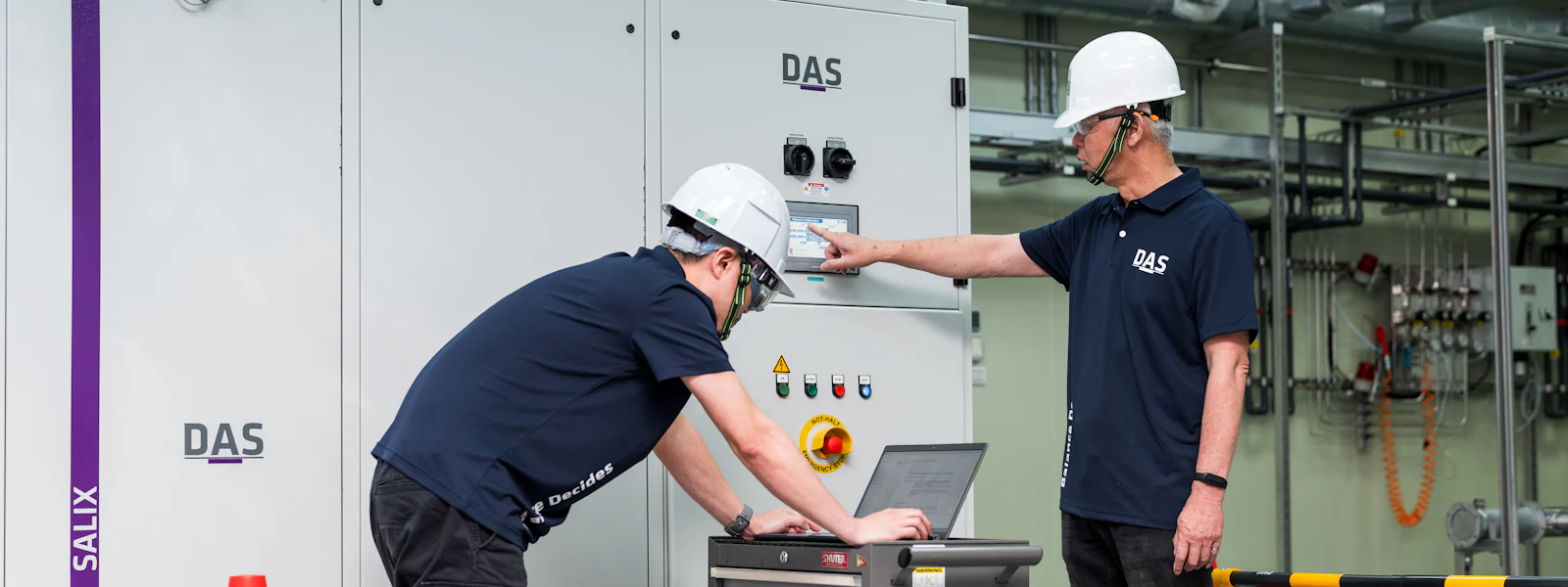 機台處理 兩位穿著深藍色制服和白色安全帽的人員在工業控制室進行設備監測,他們站在大型白色控制面板前,使用筆記型電腦檢查數據,旁邊設有安全障礙構件和反光交通錐。