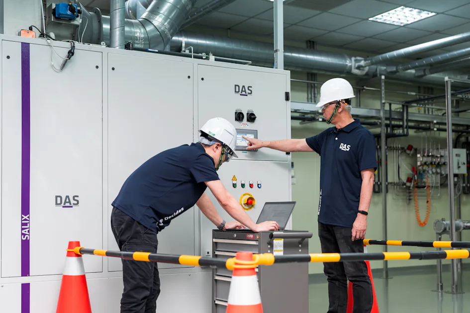 機台處理 兩位穿著深藍色制服和白色安全帽的人員在工業控制室進行設備監測,他們站在大型白色控制面板前,使用筆記型電腦檢查數據,旁邊設有安全障礙構件和反光交通錐。