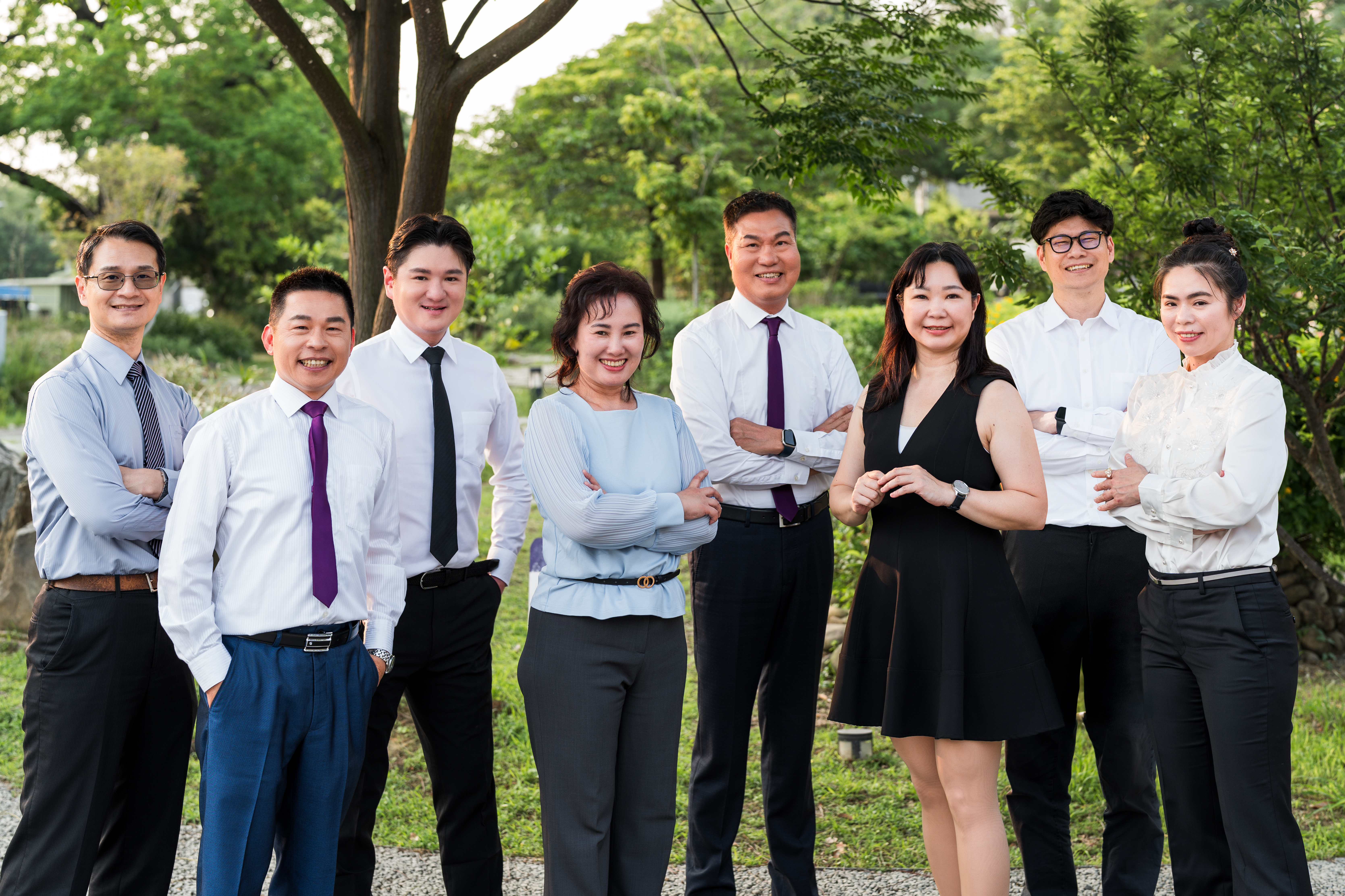 這張圖片展示了一群穿著正式商務服裝的八位人士，他們站在公園環境中，背景是綠樹和草地。