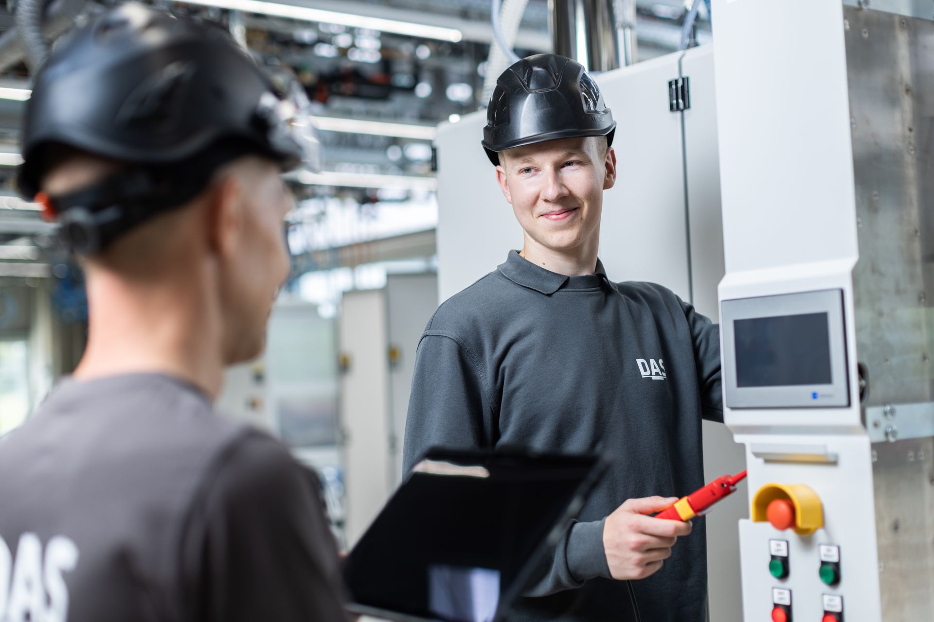 Two employees wearing black safety helmets and grey work uniforms are in a modern industrial facility. One is using a multimeter on a control panel, while the other is holding a tablet.