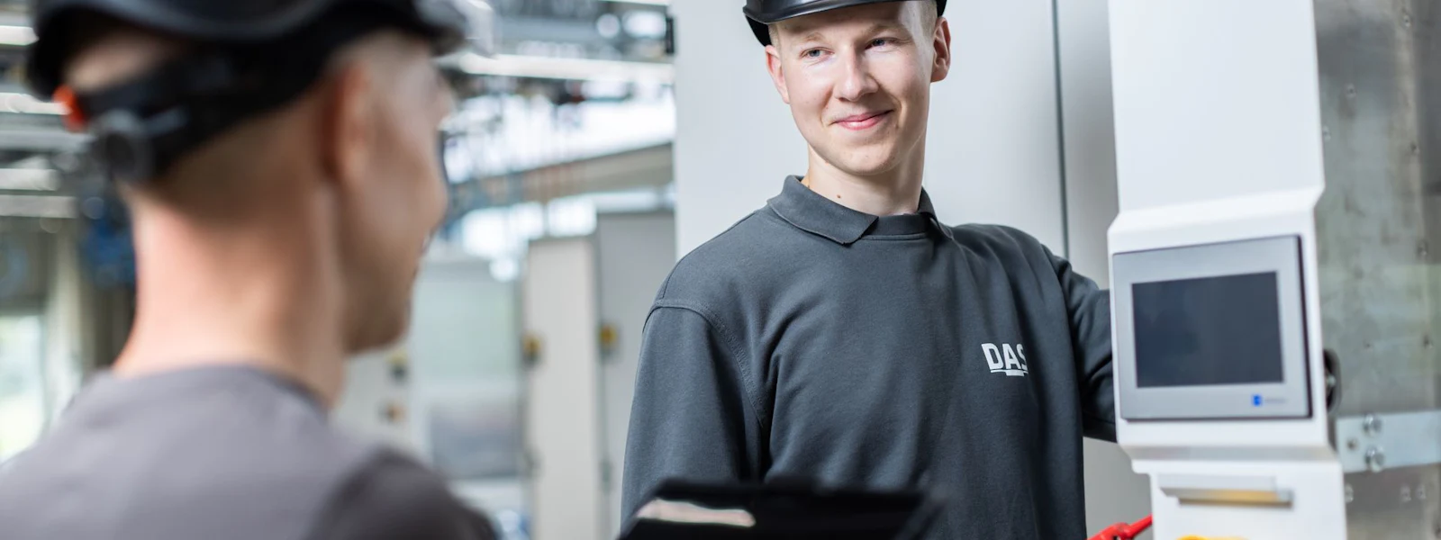 Kontrolle der Abgasreinigungsanlage Zwei Mitarbeitet mit schwarzen Schutzhelmen und grauer Arbeitskleidung Uniformen befinden sich in einer modernen Industrieanlage. Der eine benutzt ein Multimeter an einem Bedienfeld, während der andere ein Tablet in der Hand hält.