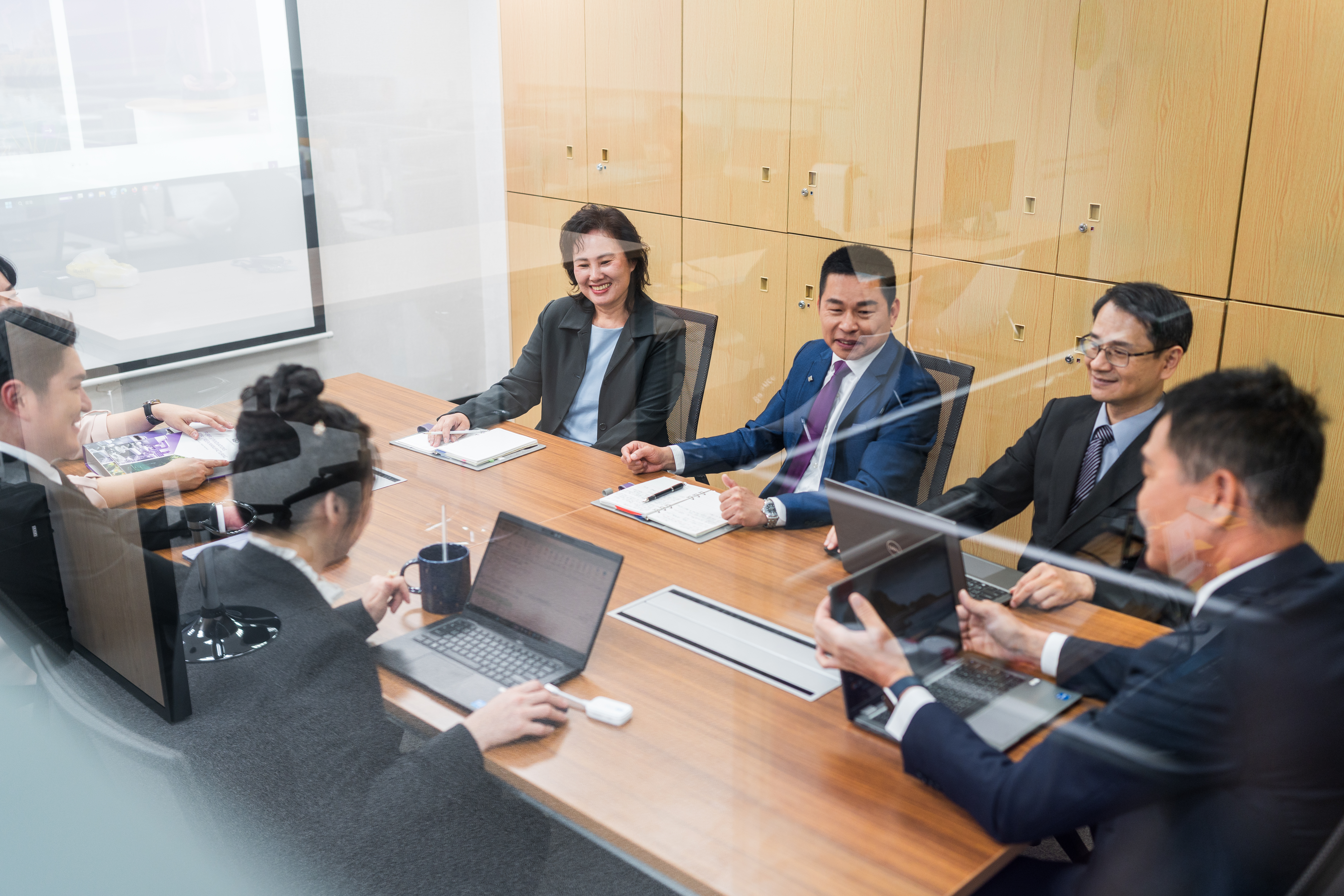 辦公室會議室內，八位商務人士圍坐在木製長桌旁，各自使用筆記本電腦進行專業討論，背景為淺色的檔案櫃。