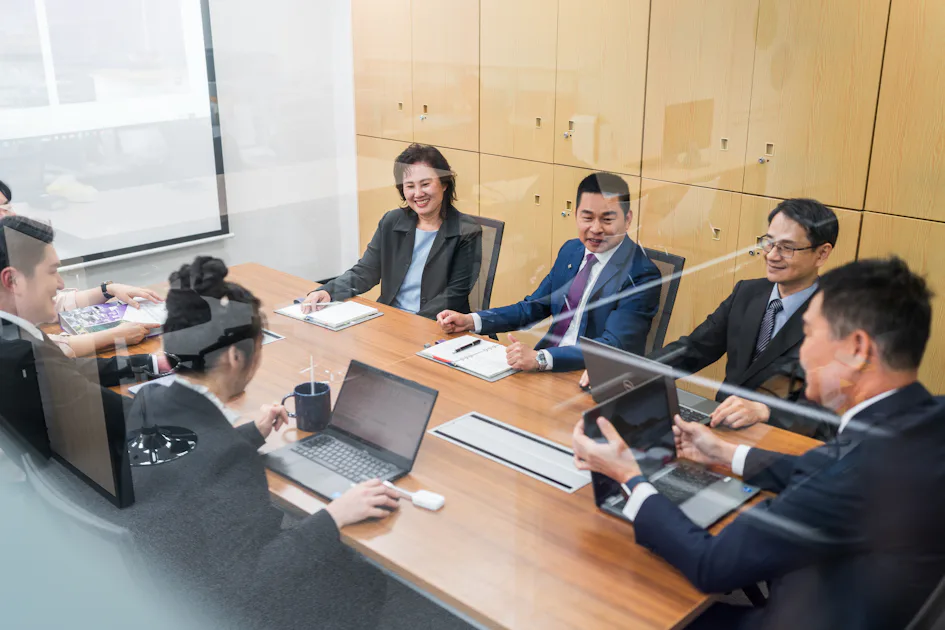 主管會議 辦公室會議室內,八位商務人士圍坐在木製長桌旁,各自使用筆記本電腦進行專業討論,背景為淺色的檔案櫃。
