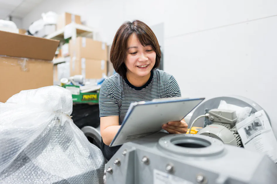 物料登記 在這張照片中,一位穿著條紋上衣的人正在工業設備附近微笑,並翻閱夾板文件,背景堆滿了紙箱和包裝材料,顯示出忙碌的工作環境。