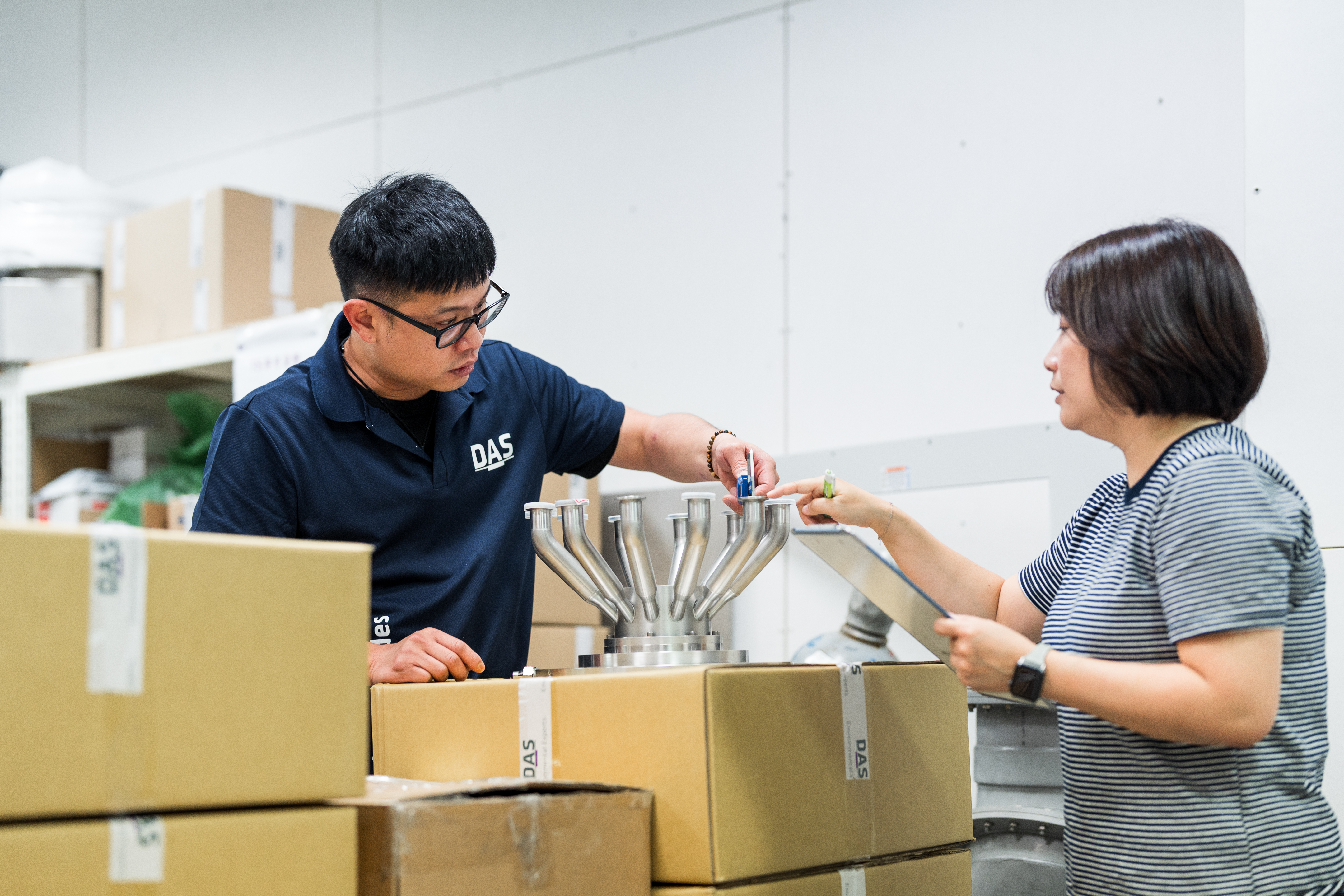 兩個人在工作場合中討論包裝箱上方的機械裝置，背景為擺滿紙箱的貨架，營造出工業環境的氛圍。