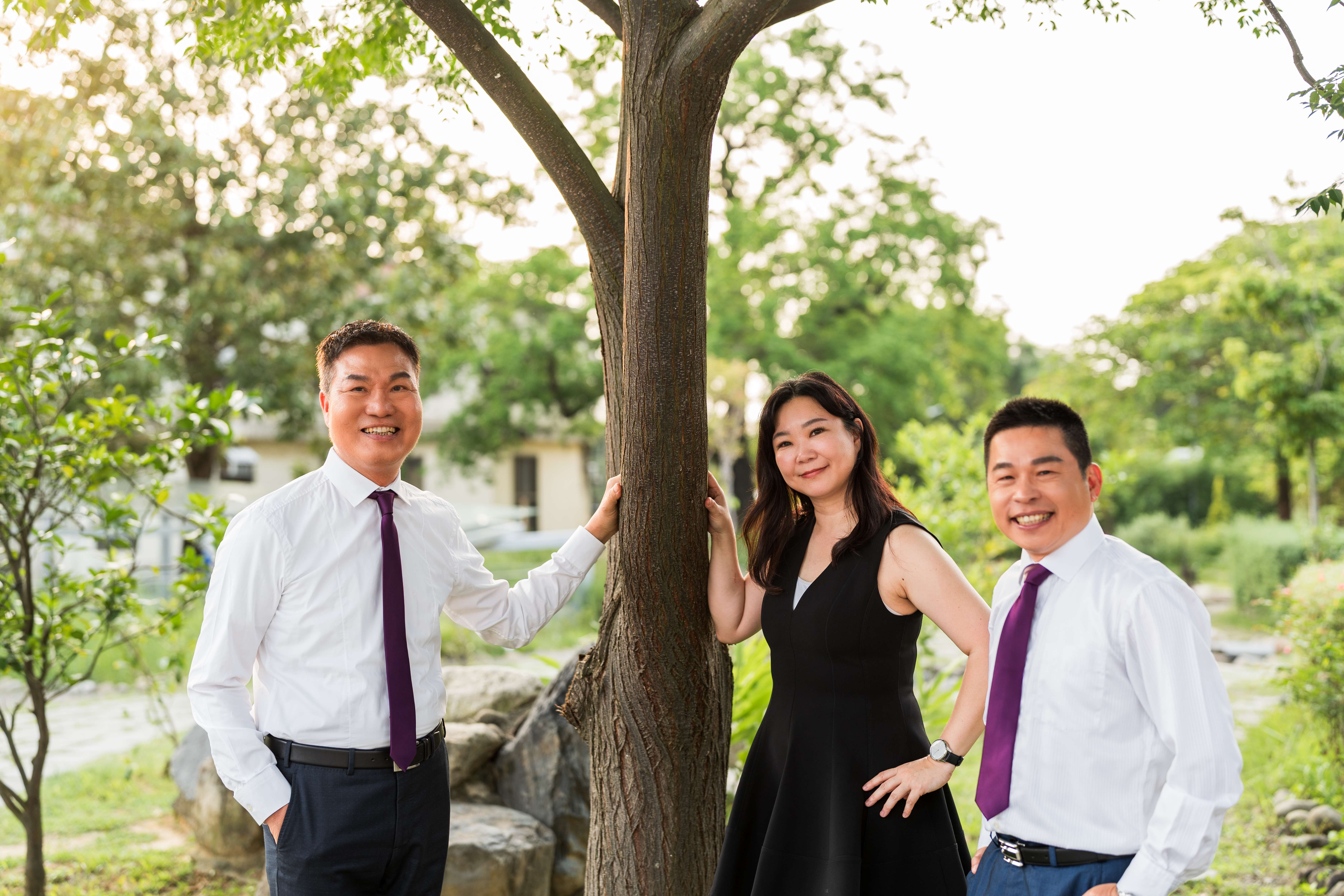 三位穿著正式服裝的人士站在樹旁微笑，背景是綠意盎然的公園景色，陽光透過樹葉灑下。