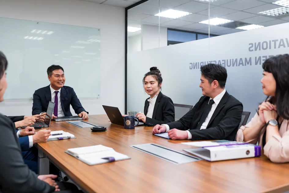 開會 六位穿著正裝的商務人士在現代會議室內開會,桌上擺放著筆記本電腦、筆記本和文件,背景有玻璃牆和白板。