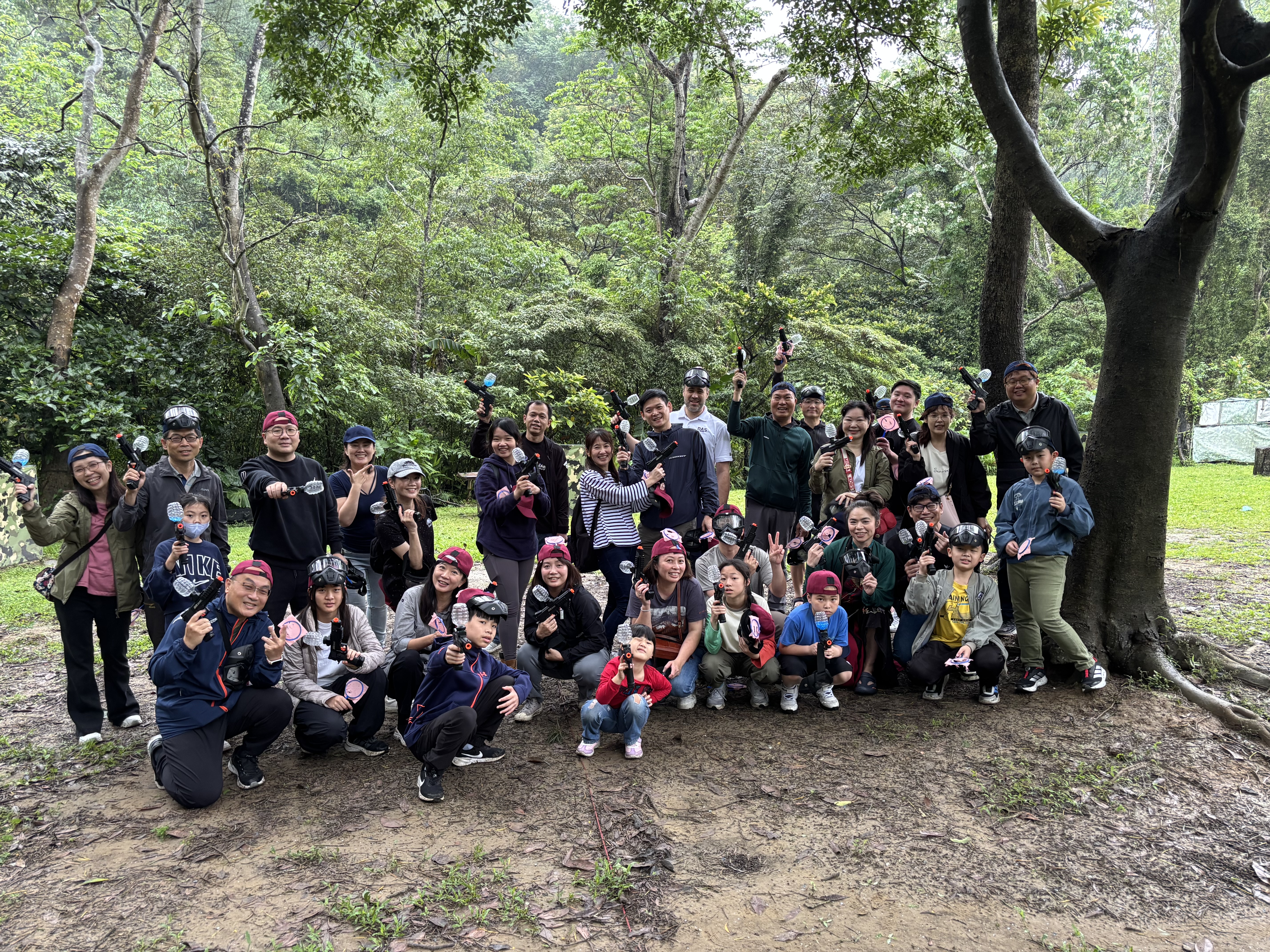 一群人在樹林中的戶外郊遊活動合影，他們站在樹下，身穿休閒服裝，展現出開心的表情。