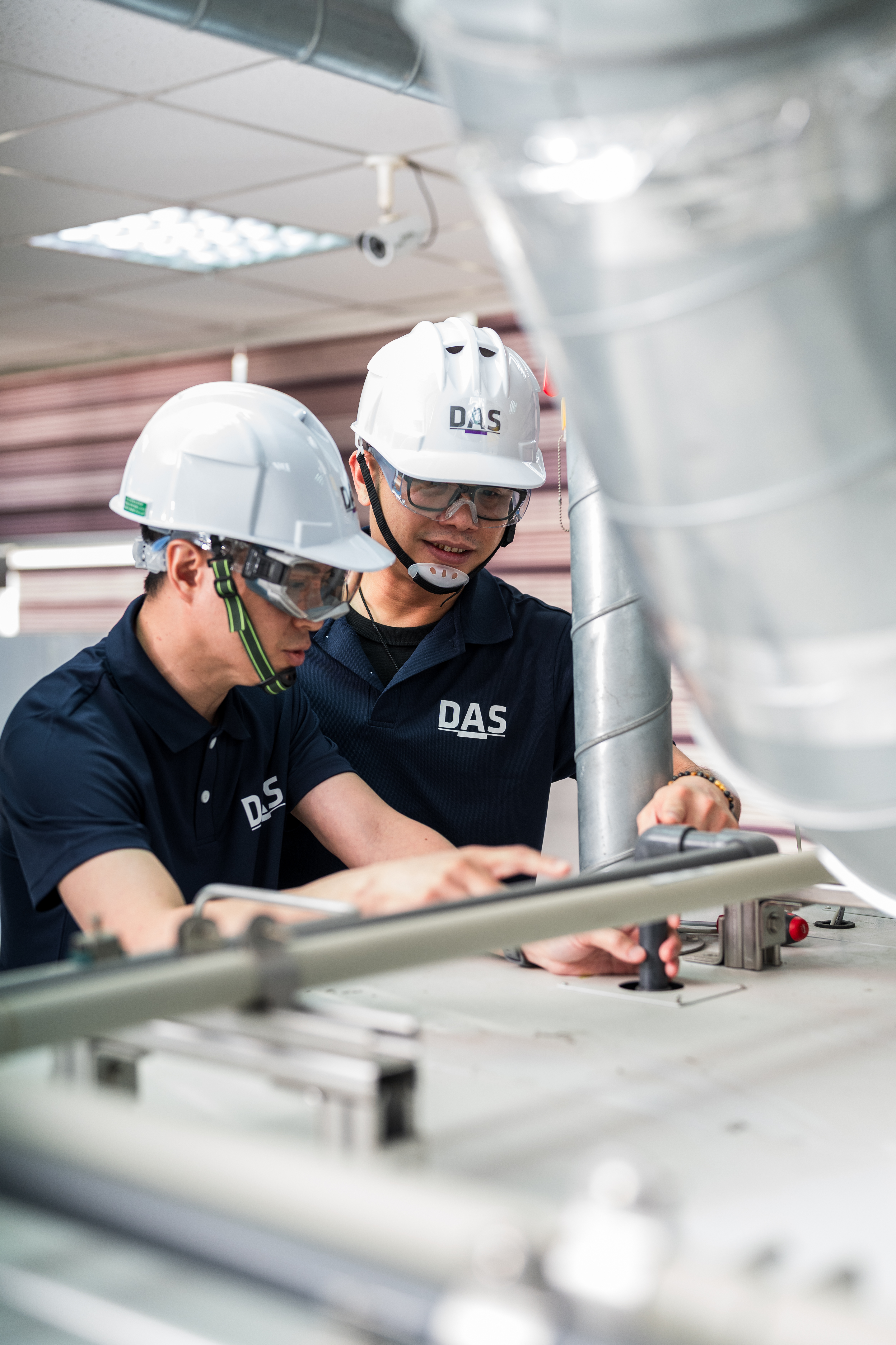 兩位戴著安全帽和護目鏡的工人，正認真地在工廠生產線上進行設備維護。