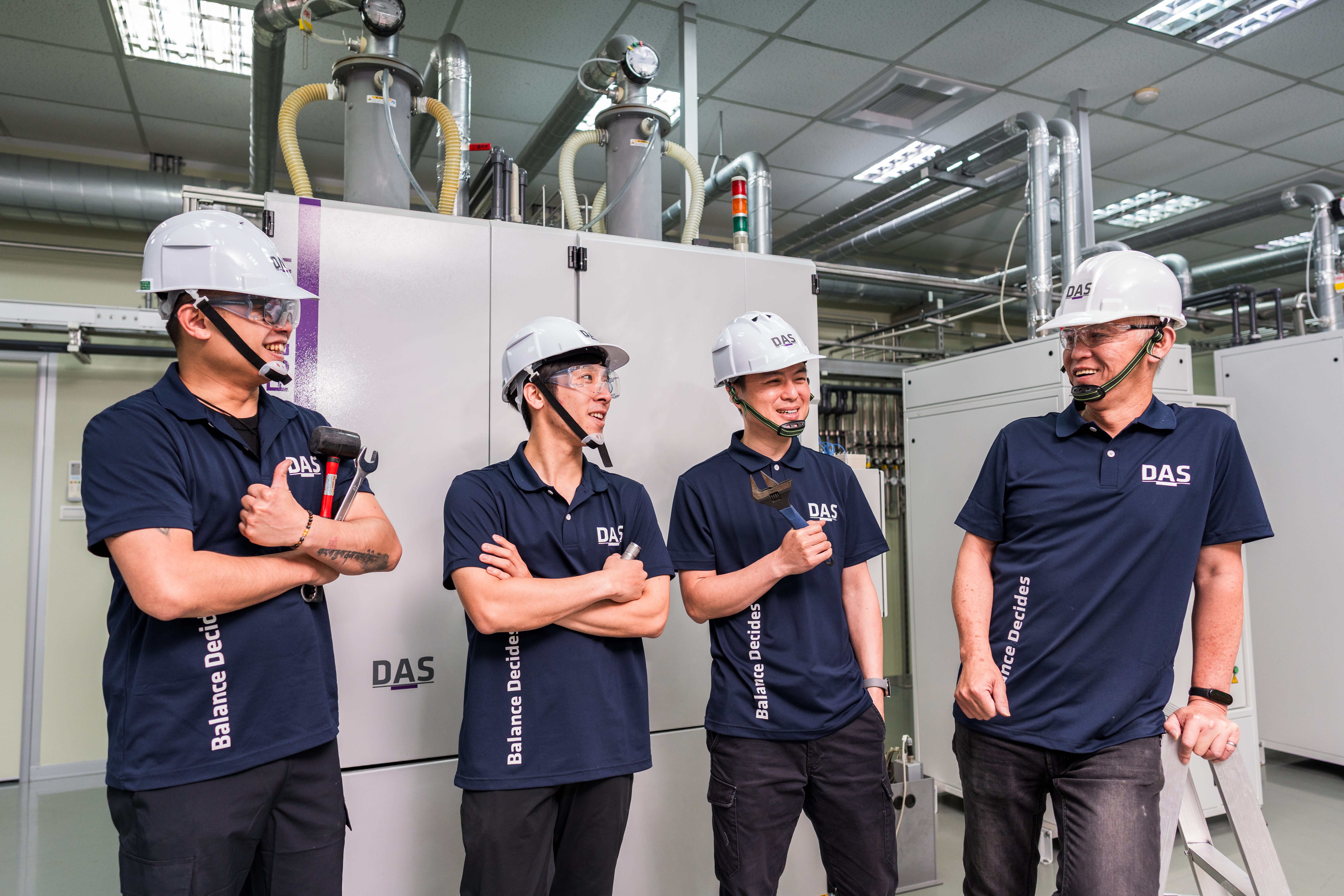 Four experts in dark blue shirts and white safety helmets stand laughing in front of technical equipment in an industrial building.