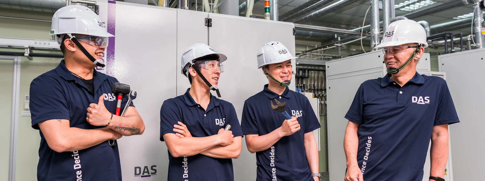 DAS Service Team Four experts in dark blue shirts and white safety helmets stand laughing in front of technical equipment in an industrial building.