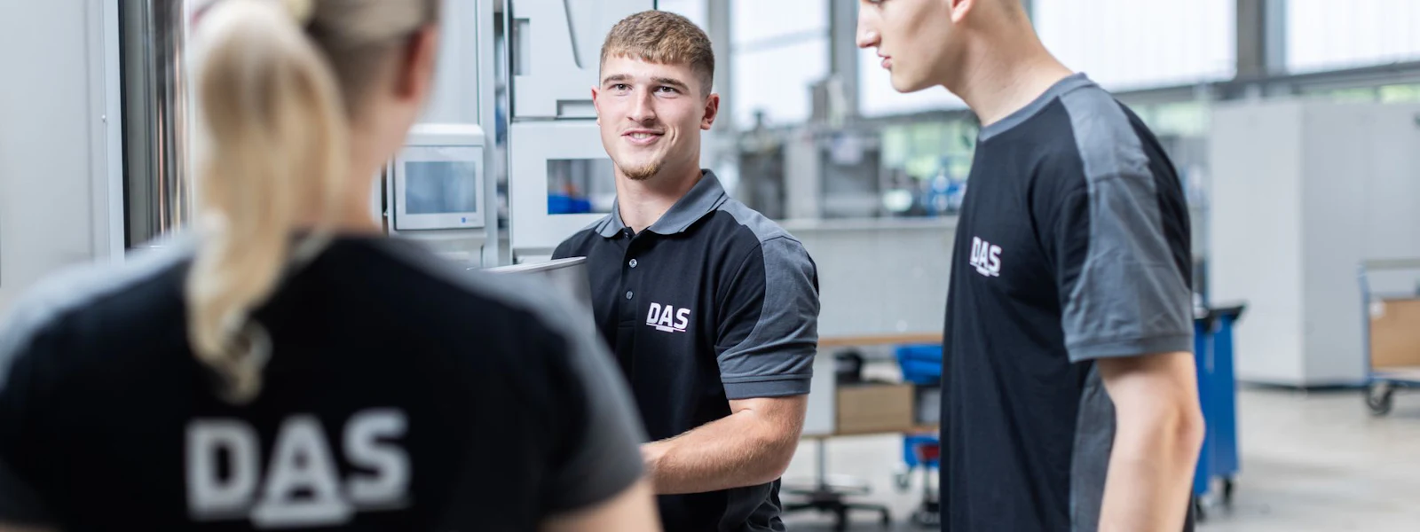 Production team in discussion Three people in work clothes are standing together in a modern, brightly lit factory hall, chatting in front of a large machine.
