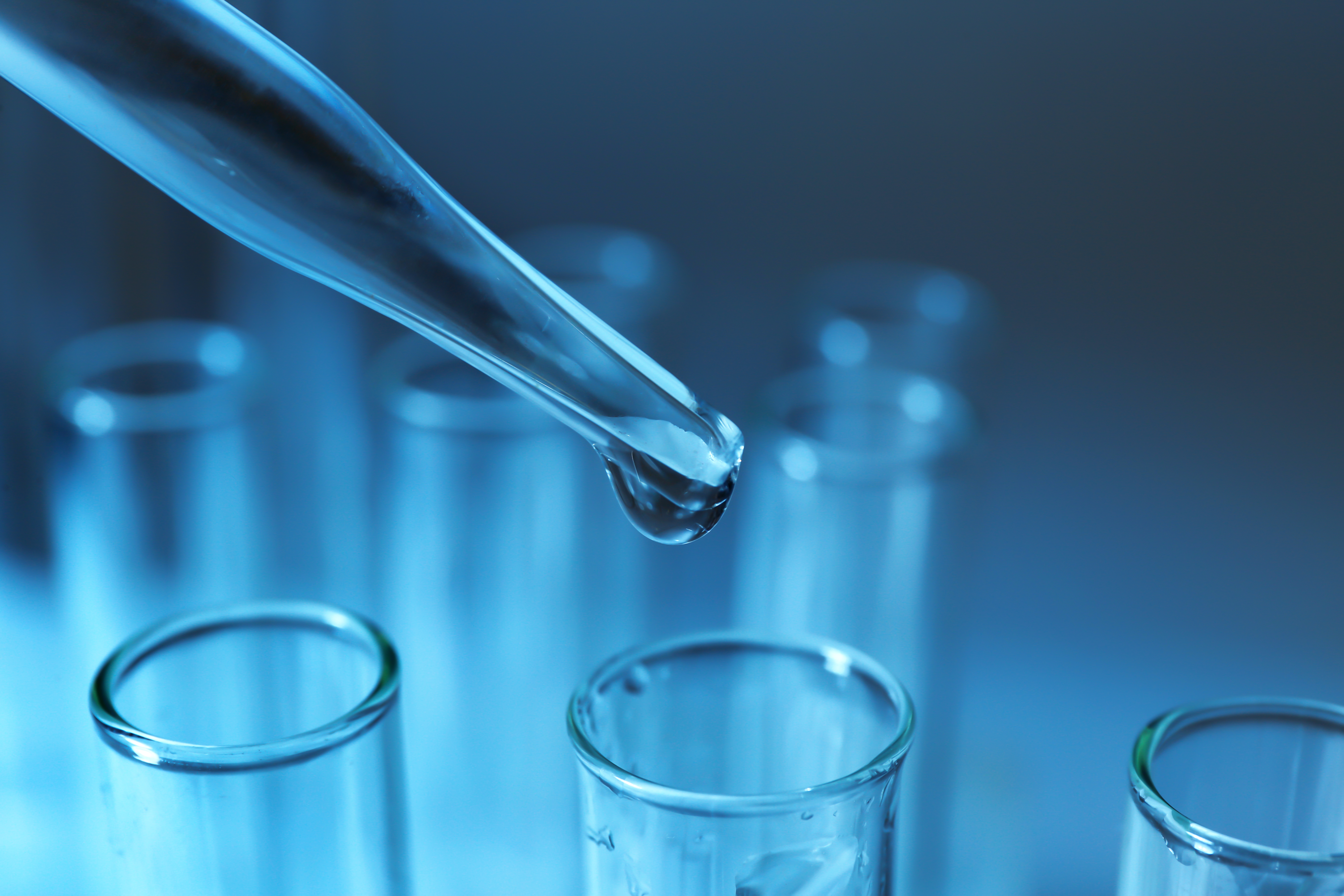 Close-up of a laboratory pipette dripping a clear liquid into a series of transparent test tubes against a cool blue background, illustrating a scientific experiment or research environment.