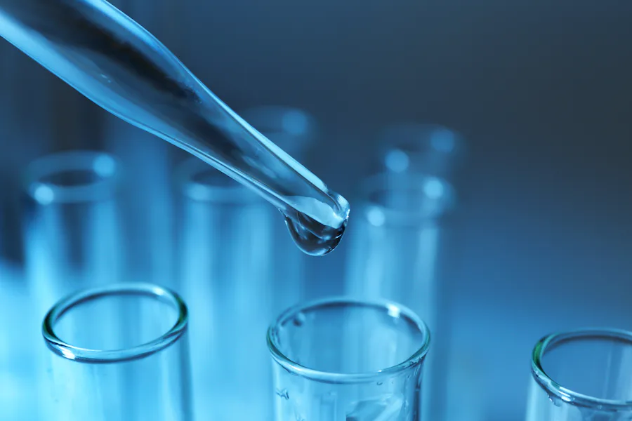 Pipette dripping sample into test tube Close-up of a laboratory pipette dripping a clear liquid into a series of transparent test tubes against a cool blue background, illustrating a scientific experiment or research environment.