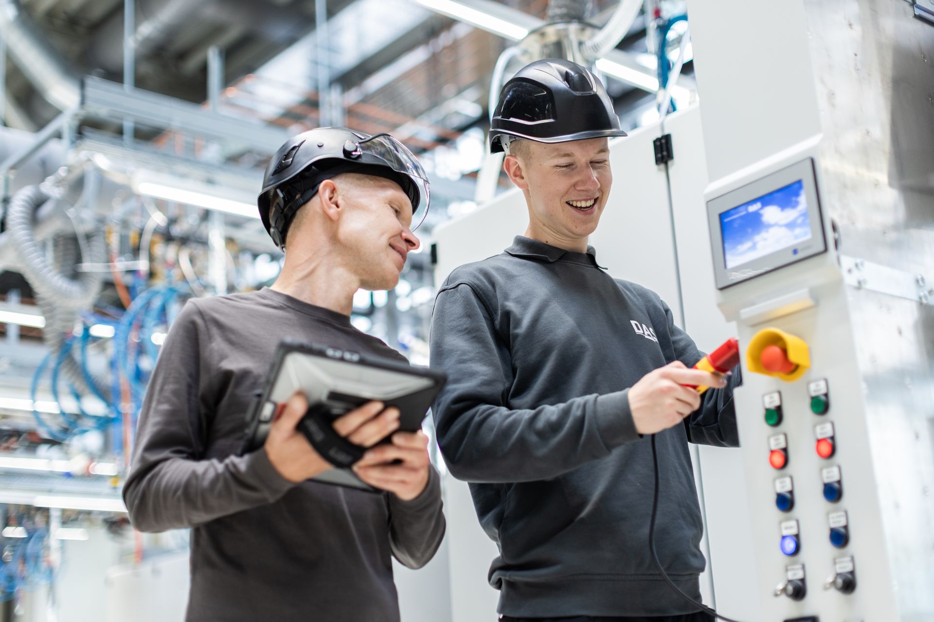 Zwei Personen in Schutzhelmen und Arbeitskleidung arbeiten zusammen in einer modernen hellen Industriehallen an einer offenen Anlage. Einer hält ein digitales Tablet, während der andere an einem Kontrollpanel mit mehreren Schaltern und einem Display arbeitet.