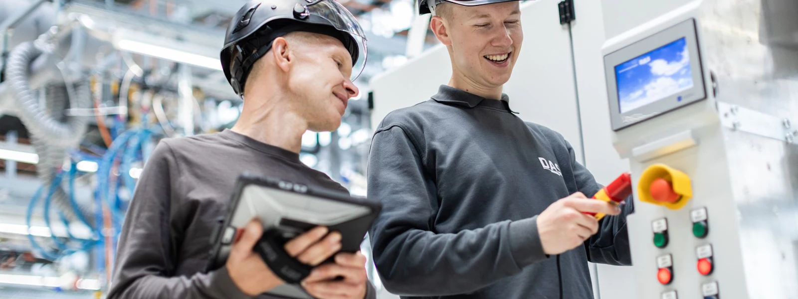 DAS Team an Anlage Zwei Personen in Schutzhelmen und Arbeitskleidung arbeiten zusammen in einer modernen hellen Industriehallen an einer offenen Anlage. Einer hält ein digitales Tablet, während der andere an einem Kontrollpanel mit mehreren Schaltern und einem Display arbeitet.