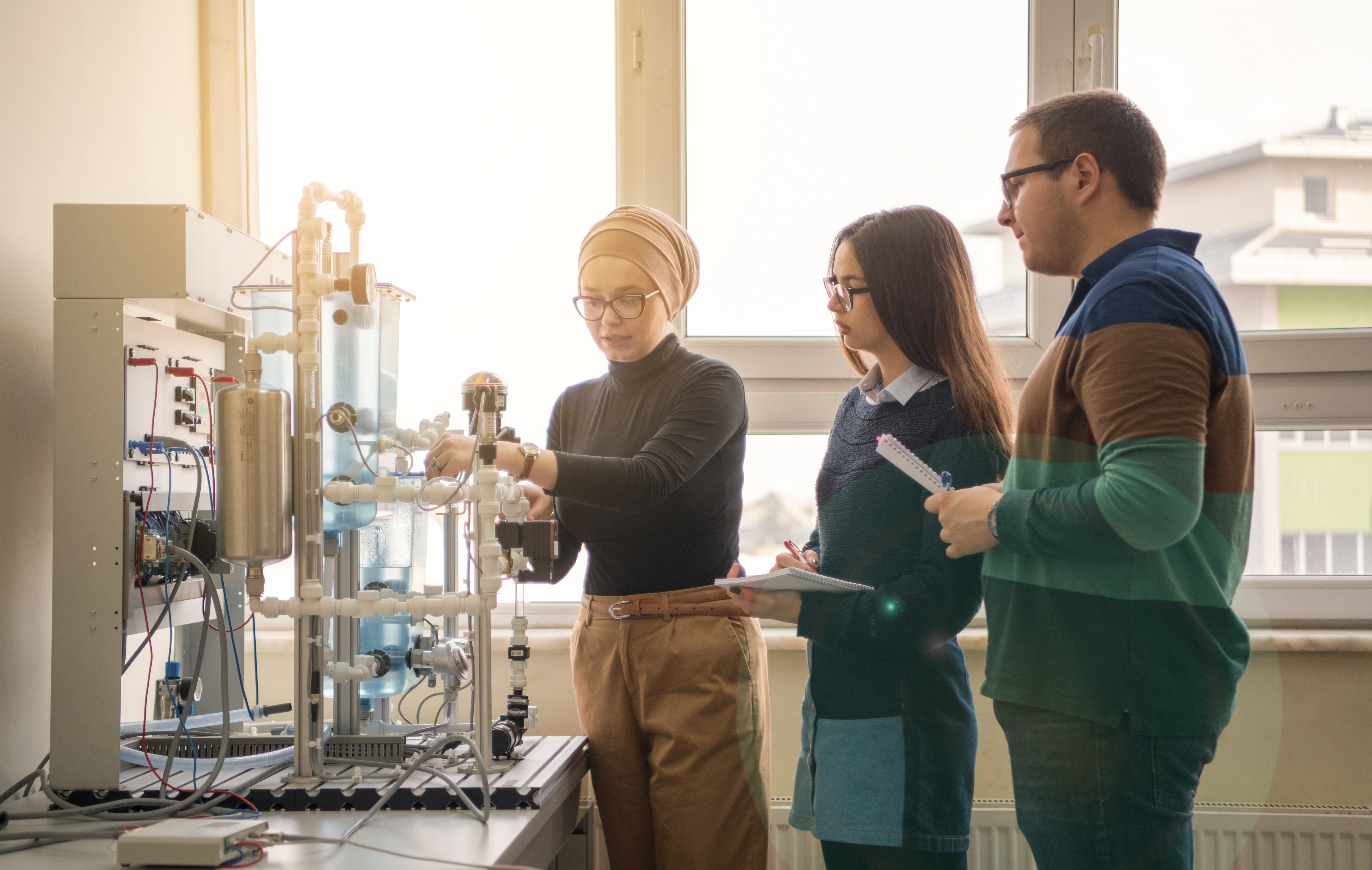 Three people are observing and discussing a mechanical apparatus in a bright laboratory setting, demonstrating teamwork and collaboration in scientific research and engineering.