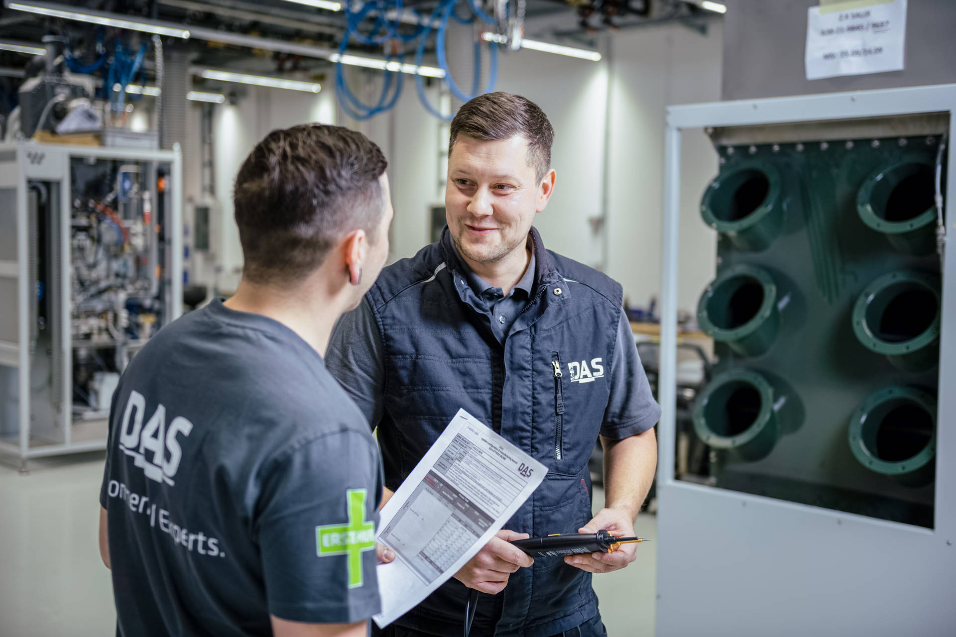 Two men in an industrial production hall discuss with a plan in their hands while looking at a large plant with several large supply lines.