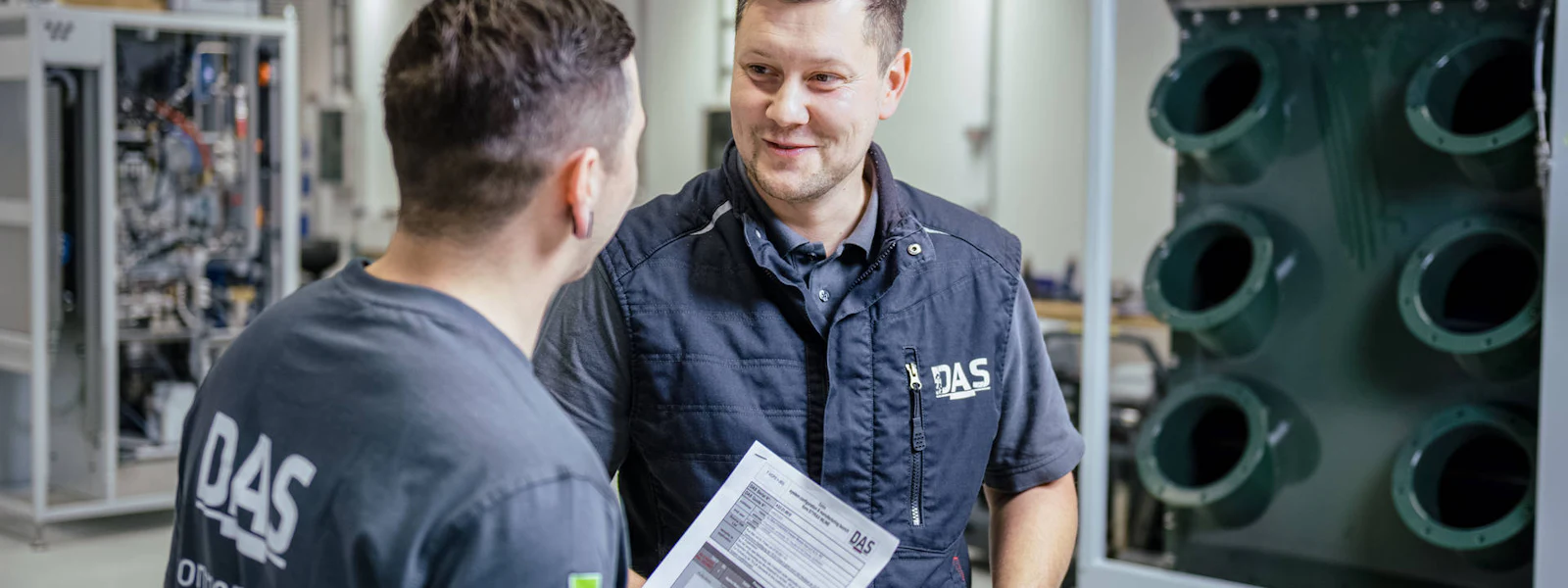 Fertigung SALIX Zwei Männer in einer industriellen Fertigungshalle diskutieren mit einem Plan in der Hand, während sie eine große Anlage mit mehreren großen Zuleitungen betrachten.
