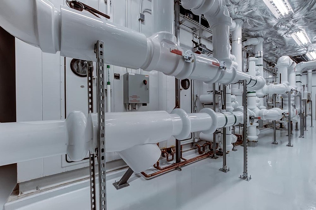 A modern industrial piping system with shiny white pipes and metal brackets runs through a room with grey flooring.