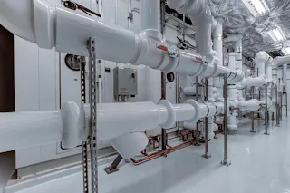 Piping System in a fab A modern industrial piping system with shiny white pipes and metal brackets runs through a room with grey flooring.