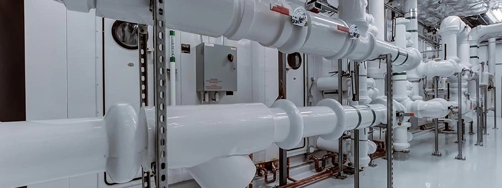 Piping System in a fab A modern industrial piping system with shiny white pipes and metal brackets runs through a room with grey flooring.